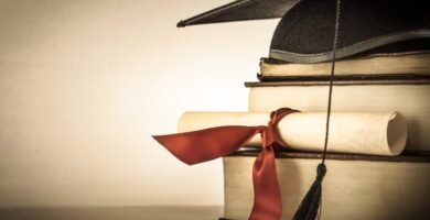 Zdjęcie stosu książek pod czapką i dyplomem ukończenia szkoły na brązowym tle.